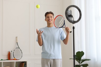 Photo of Smiling sports blogger holding tennis racket and ball while streaming online fitness lesson at home