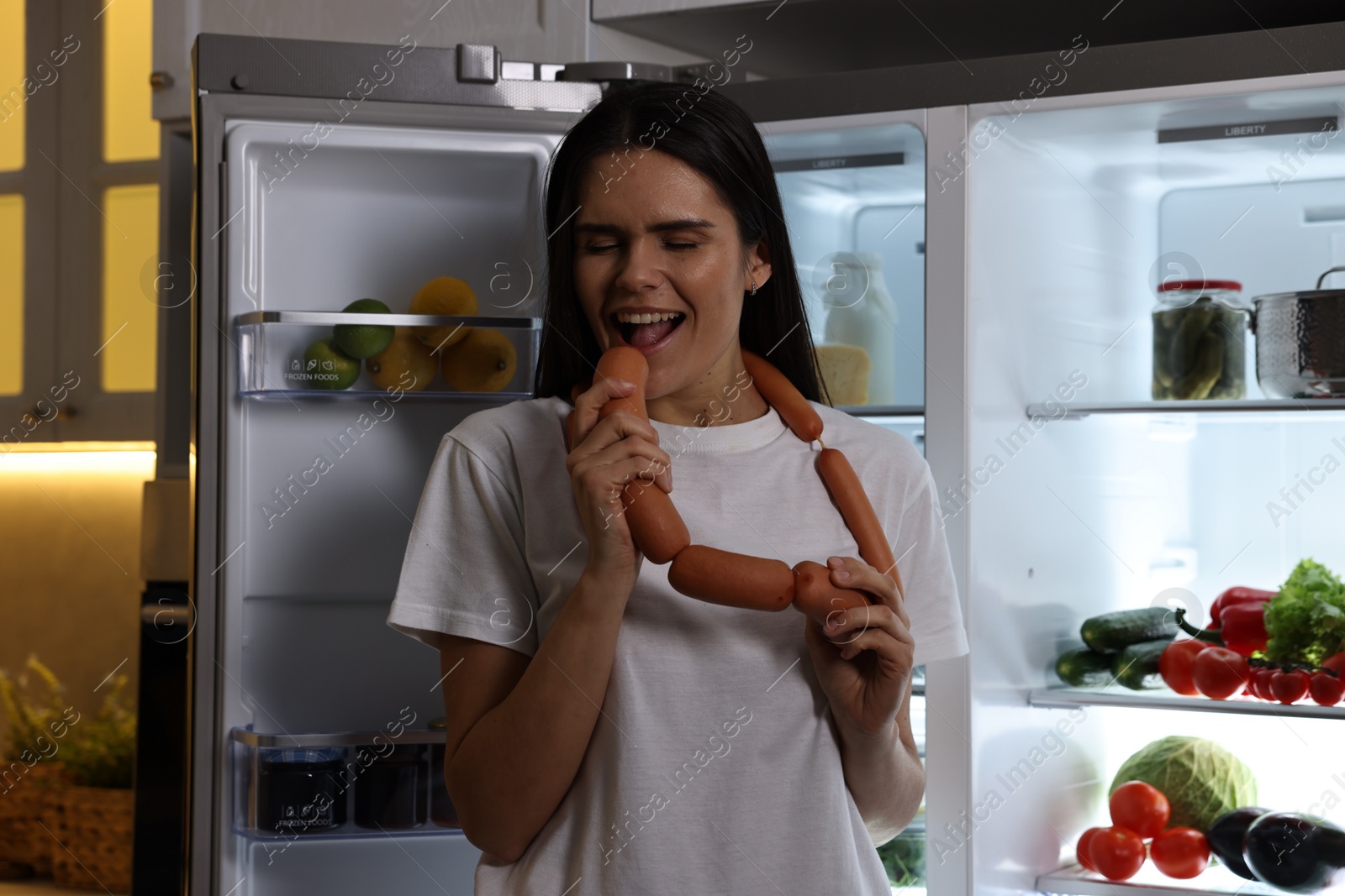 Photo of Young woman eating sausages near modern refrigerator in kitchen at night