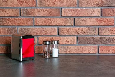 Napkin holder with salt and pepper shakers on table near brick wall. Space for text