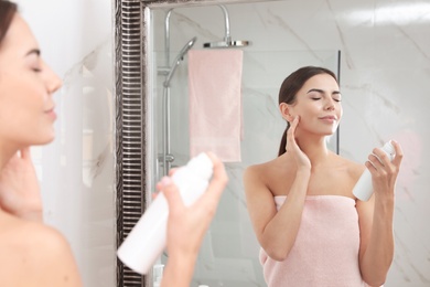 Young woman applying thermal water on face near mirror in bathroom. Cosmetic product