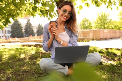 Happy young woman with paper cup of coffee and laptop sitting on green grass in park