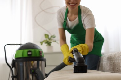Photo of Professional janitor in uniform vacuuming furniture indoors, closeup