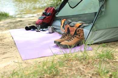 Photo of Boots and camping equipment near tent on riverbank. Space for text