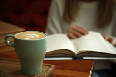 Woman with coffee reading book indoors, focus on cup