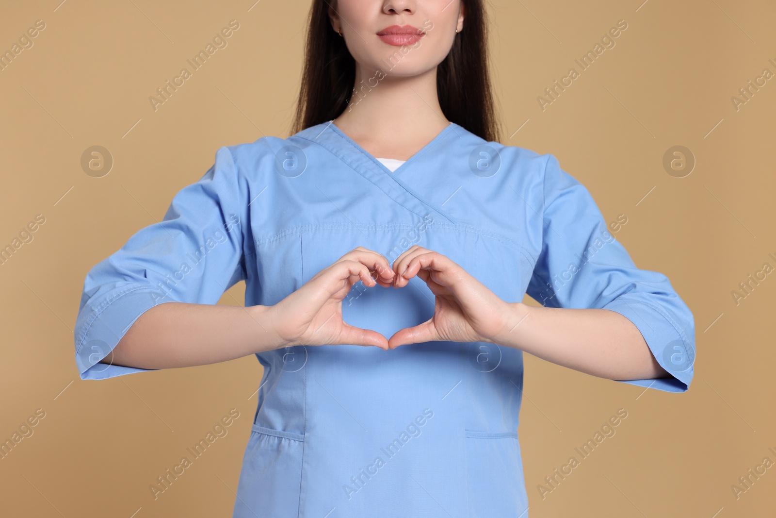 Photo of Nurse in medical uniform making heart with hands on light brown background, closeup