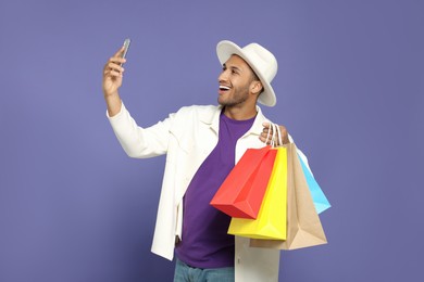 Photo of Happy African American man in hat with shopping bags taking selfie on purple background