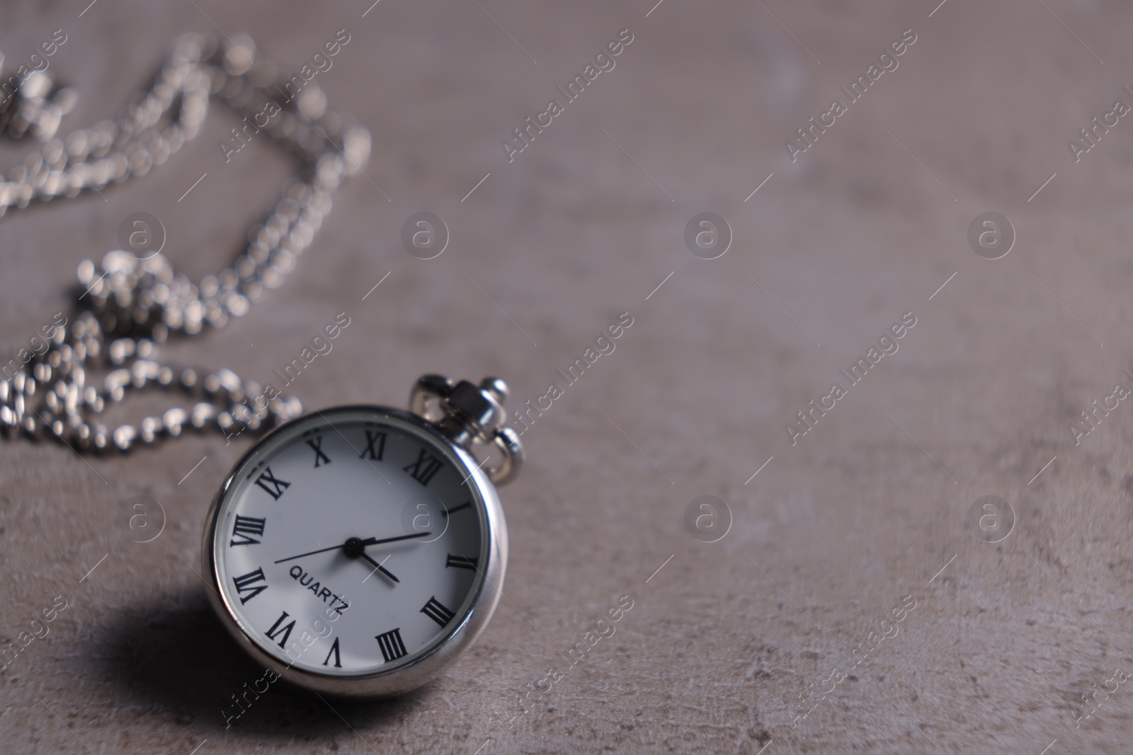 Photo of Silver pocket clock with chain on grey textured table, closeup. Space for text