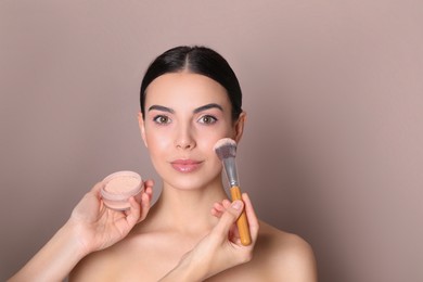 Professional makeup artist applying powder onto beautiful young woman's face with brush on dusty rose background