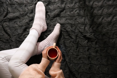 Woman in warm socks relaxing with cup of hot drink on knitted plaid, top view. Space for text