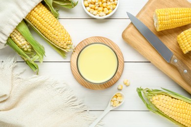Photo of Tasty fresh corn milk in glass and cobs on white wooden table, flat lay