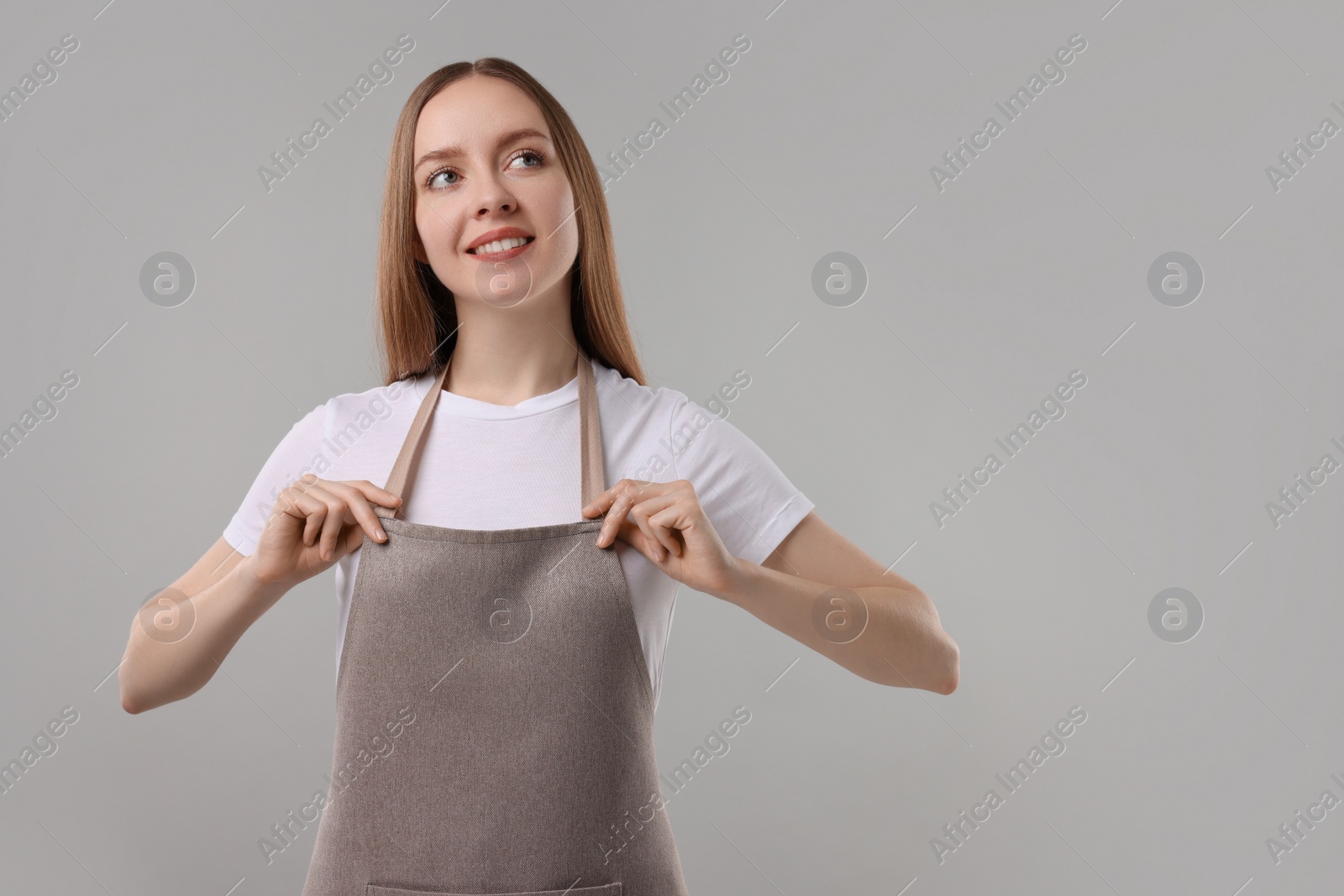 Photo of Beautiful young woman wearing kitchen apron on grey background. Mockup for design