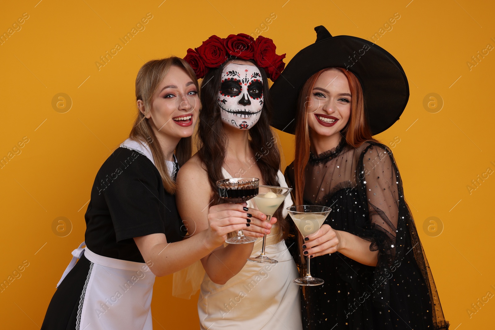 Photo of Women in scary costumes with glasses of cocktails on orange background. Halloween celebration