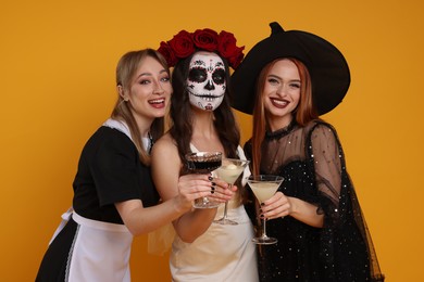 Photo of Women in scary costumes with glasses of cocktails on orange background. Halloween celebration