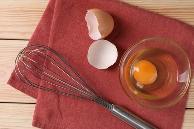 Photo of Metal whisk, raw eggs in bowl and shells on wooden table, flat lay