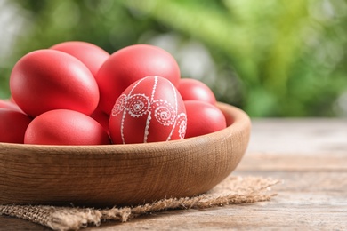 Photo of Wooden bowl with red painted Easter eggs on table against blurred background, space for text