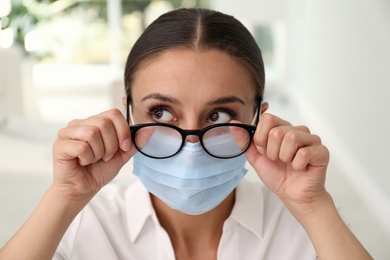 Woman wiping foggy glasses caused by wearing medical mask indoors, closeup