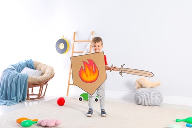 Photo of Cute little boy playing with cardboard sword and shield at home
