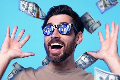 Image of Excited man under money shower on light blue background. Abbreviation NFT reflecting in sunglasses