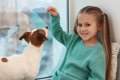 Cute girl training her dog on window sill indoors. Adorable pet