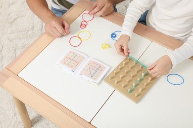 Photo of Motor skills development. Father helping his daughter to play with geoboard and rubber bands at white table, above view