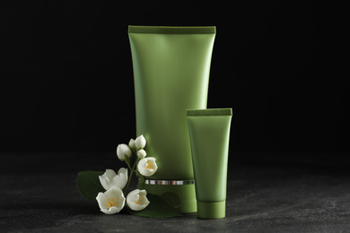 Photo of Tubes of cosmetic products and flowers on grey stone table against black background
