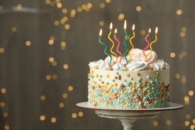 Beautiful birthday cake with burning candles on stand against festive lights. Space for text