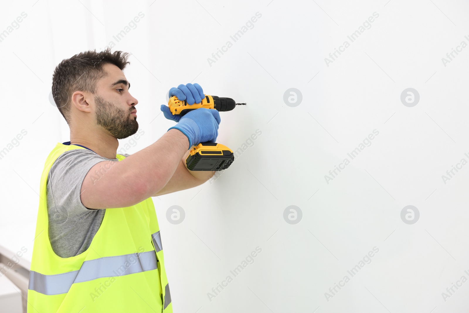 Photo of Young worker in uniform using electric drill indoors. Space for text