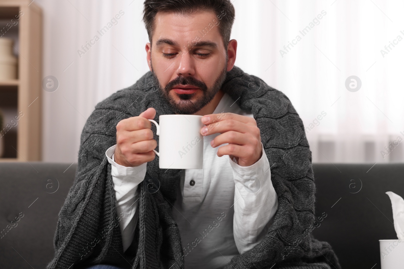 Photo of Sick man wrapped in blanket with cup of drink on sofa at home. Cold symptoms