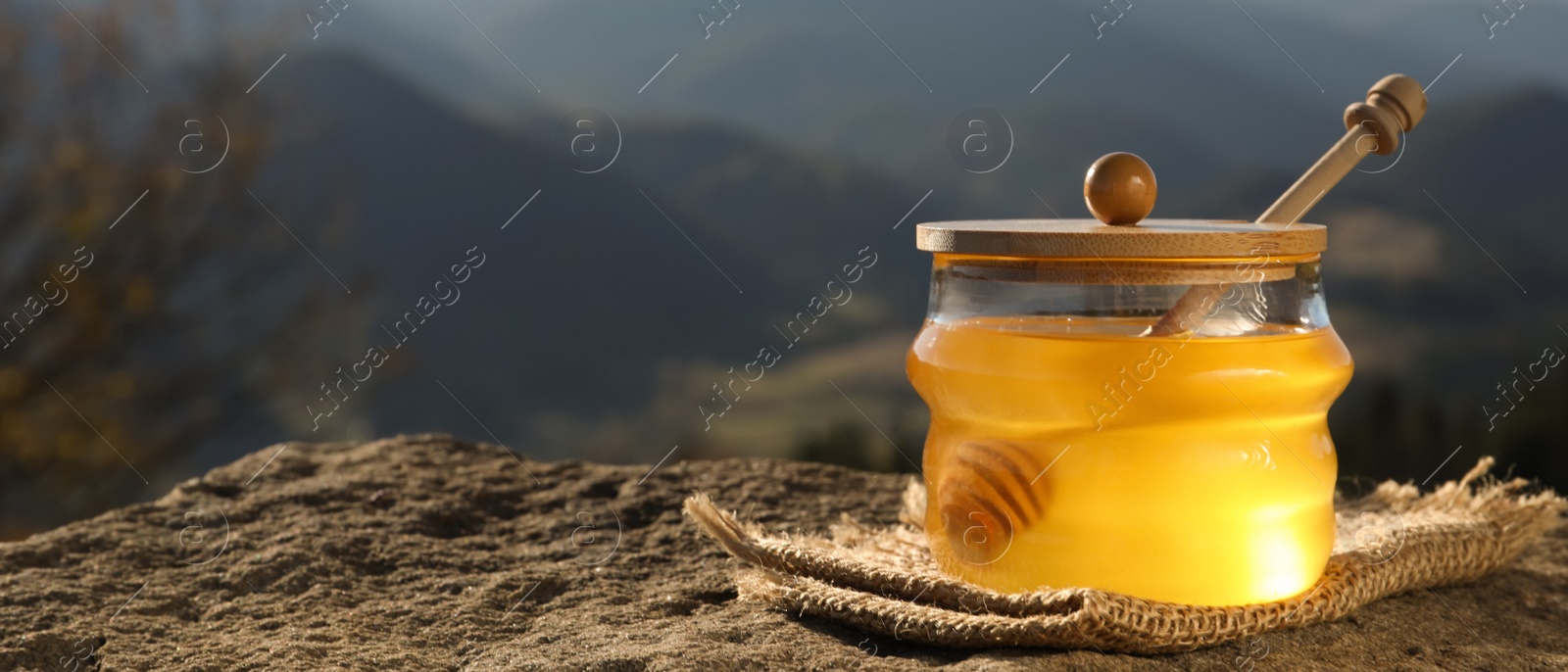 Image of Fresh aromatic honey in glass jar on rock against mountain landscape, space for text. Banner design