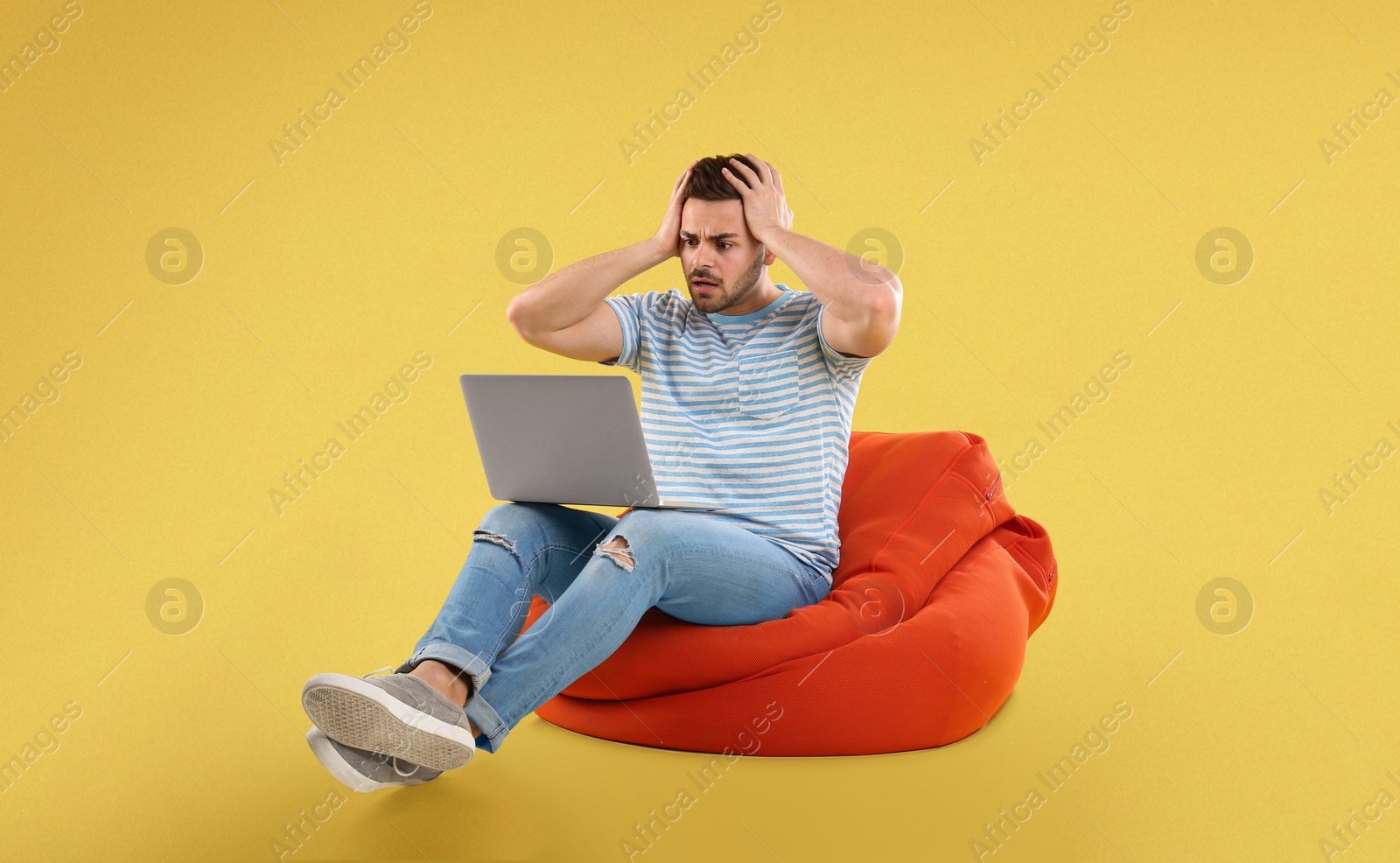 Image of Worried young man with laptop on bean bag chair, yellow background