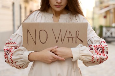 Photo of Sad woman in embroidered dress holding poster No War on city street, closeup
