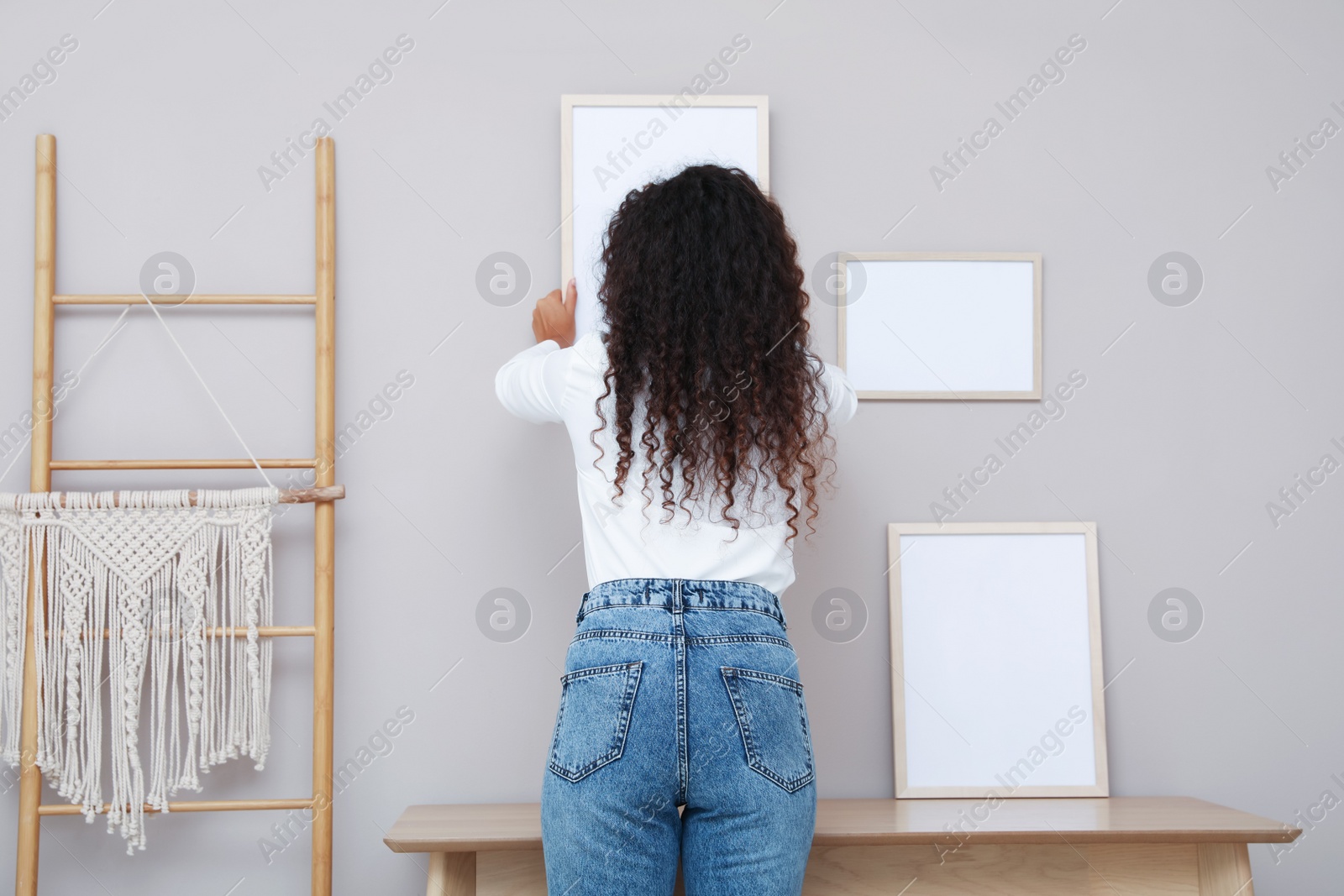 Photo of Woman hanging empty frame on pale rose wall over table in room, back view