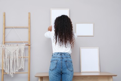 Photo of Woman hanging empty frame on pale rose wall over table in room, back view