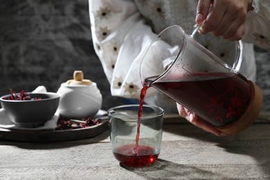 Woman pouring freshly brewed hibiscus tea from teapot into glass at wooden table, closeup