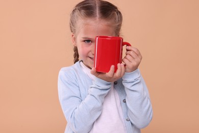 Photo of Happy girl covering eye with red ceramic mug on beige background