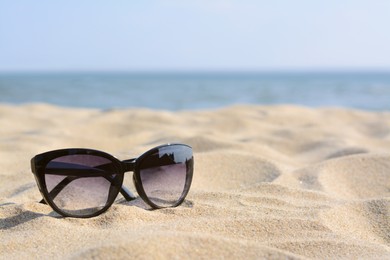 Stylish sunglasses on sandy beach near sea, space for text