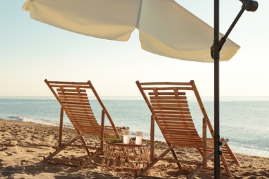 Wooden deck chairs, outdoor umbrella and table with cocktails on sandy beach. Summer vacation