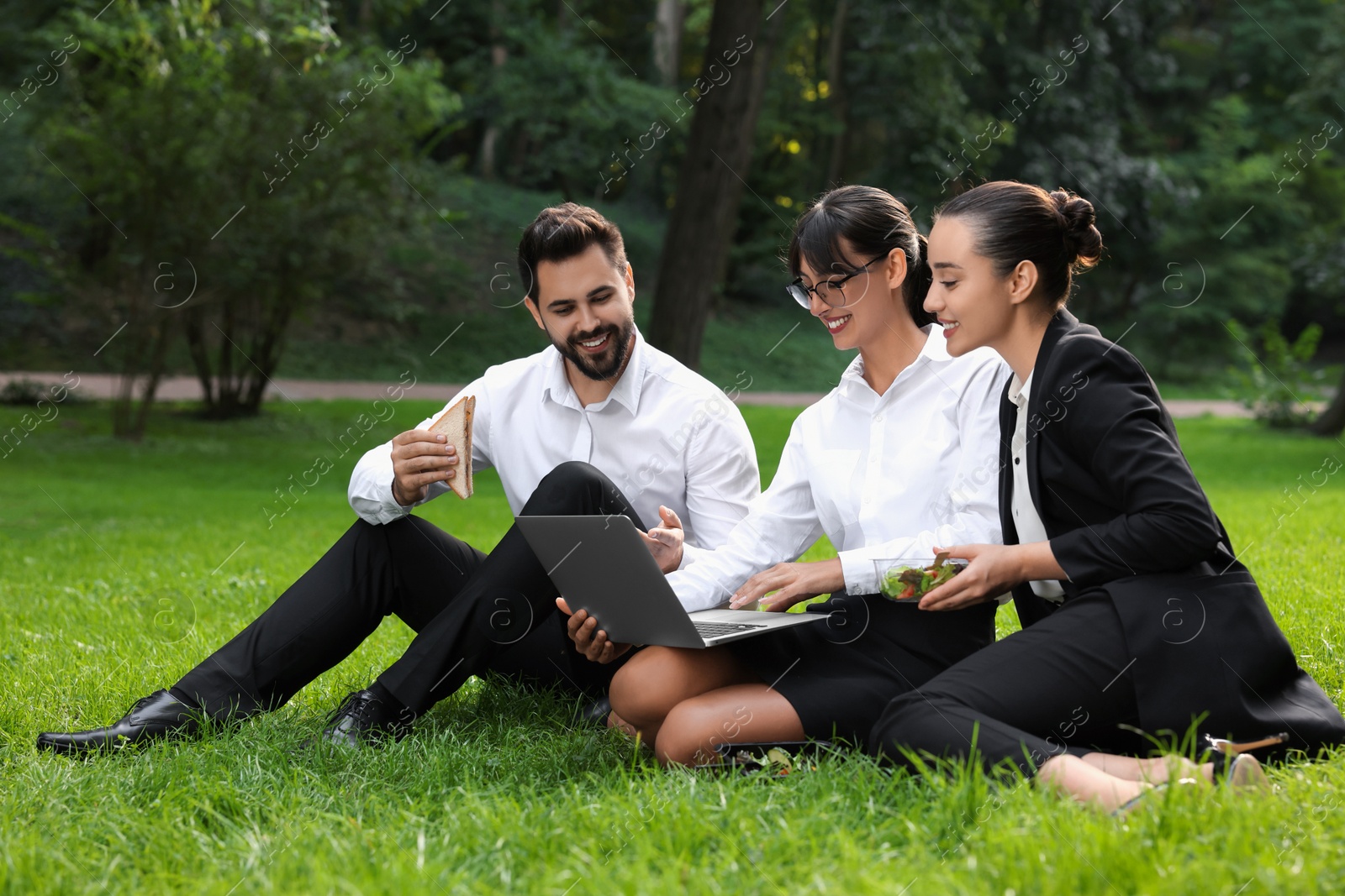 Photo of Happy colleagues with laptop having business lunch on green grass in park