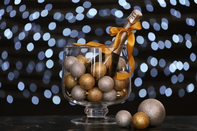 Photo of Bottle of champagne and Christmas balls in glass vase on table. New year composition against blurred lights