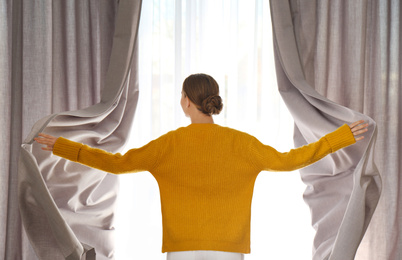 Photo of Woman opening window curtains at home in morning, back view