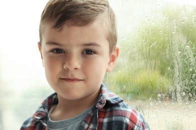 Photo of Cute little boy near window indoors, space for text. Rainy day