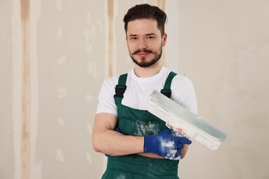 Happy worker in uniform with putty knife near plastered wall indoors