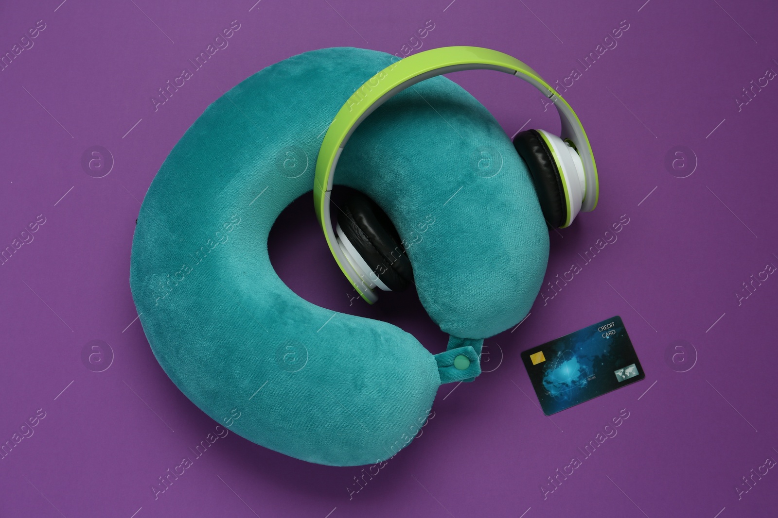 Photo of Turquoise travel pillow, headphones and credit card on purple background, flat lay