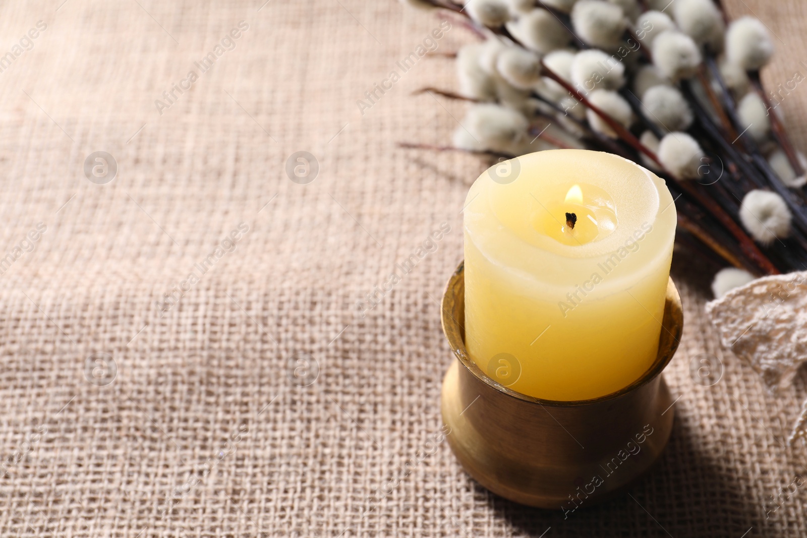 Photo of Burning church candle and willow branches on beige cloth, closeup. Space for text