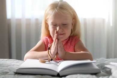 Photo of Cute little girl with beads praying over Bible in bedroom