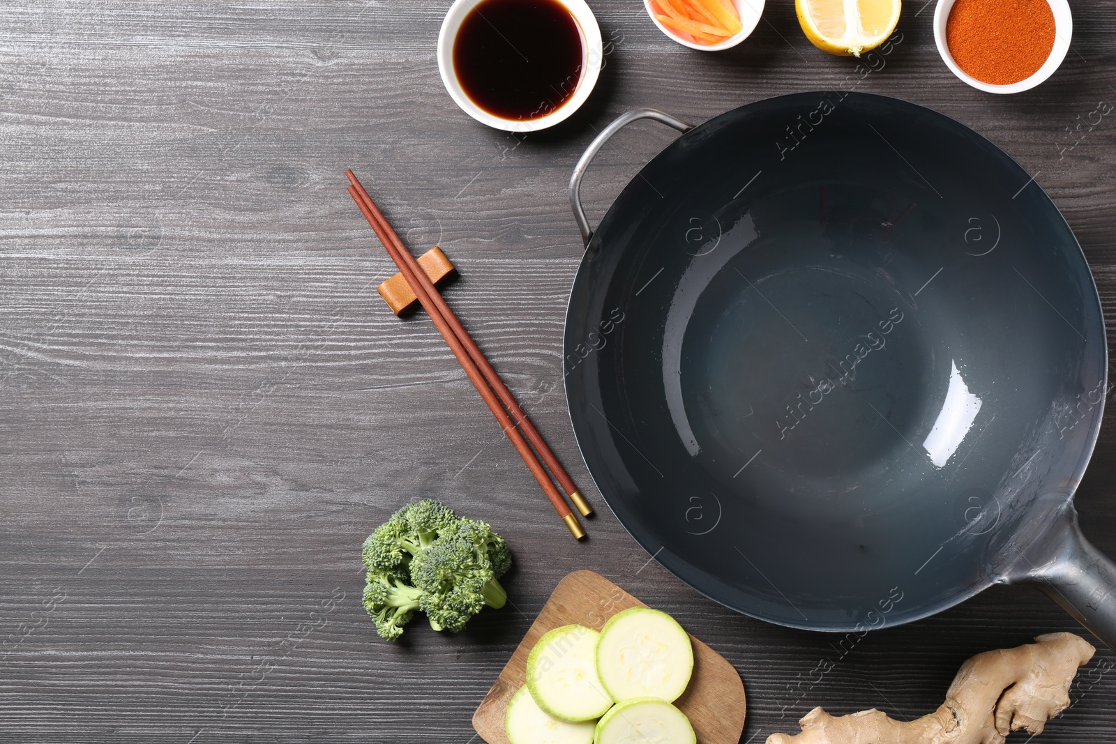 Photo of Empty iron wok, chopsticks and ingredients on grey wooden table, flat lay. Space for text