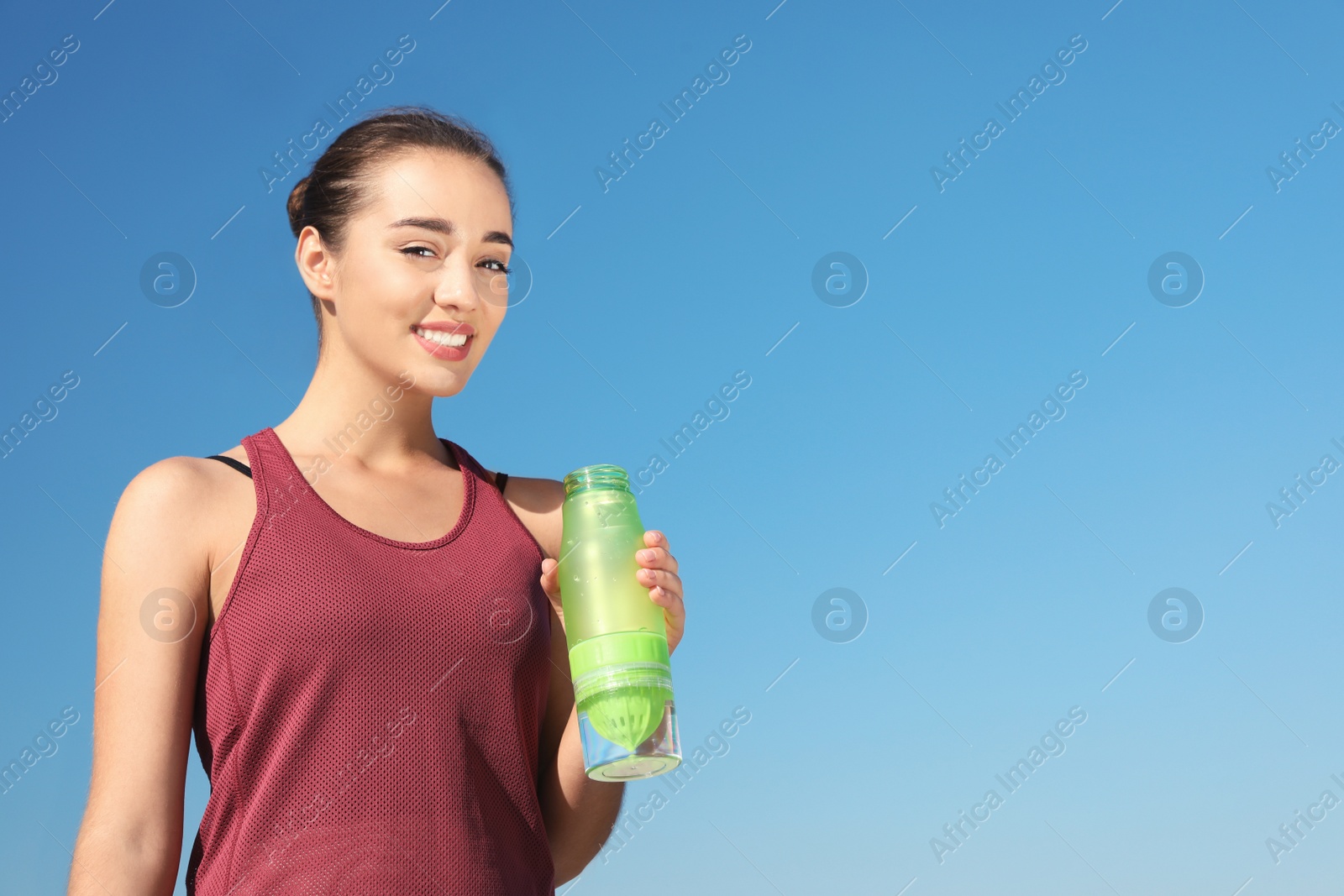 Photo of Young sporty woman holding bottle of water against blue sky on sunny day. Space for text