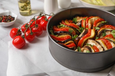 Photo of Delicious ratatouille in round baking pan and ingredients on white table
