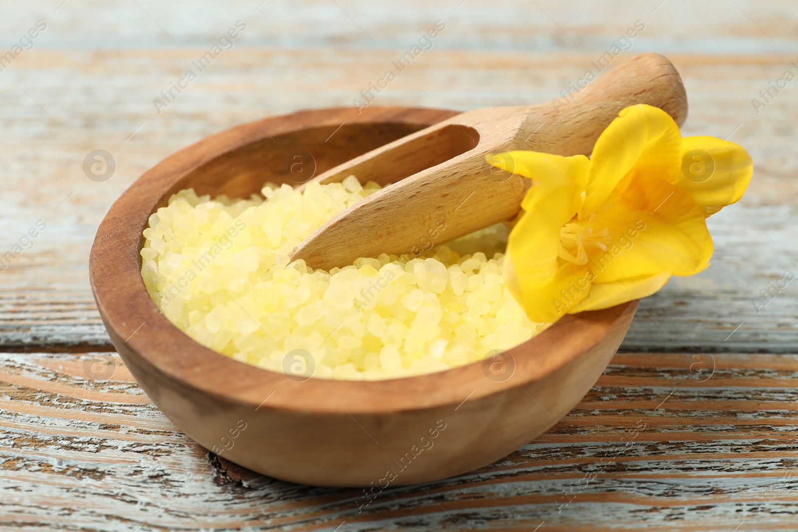 Photo of Yellow sea salt in bowl, scoop and flower on wooden table, closeup