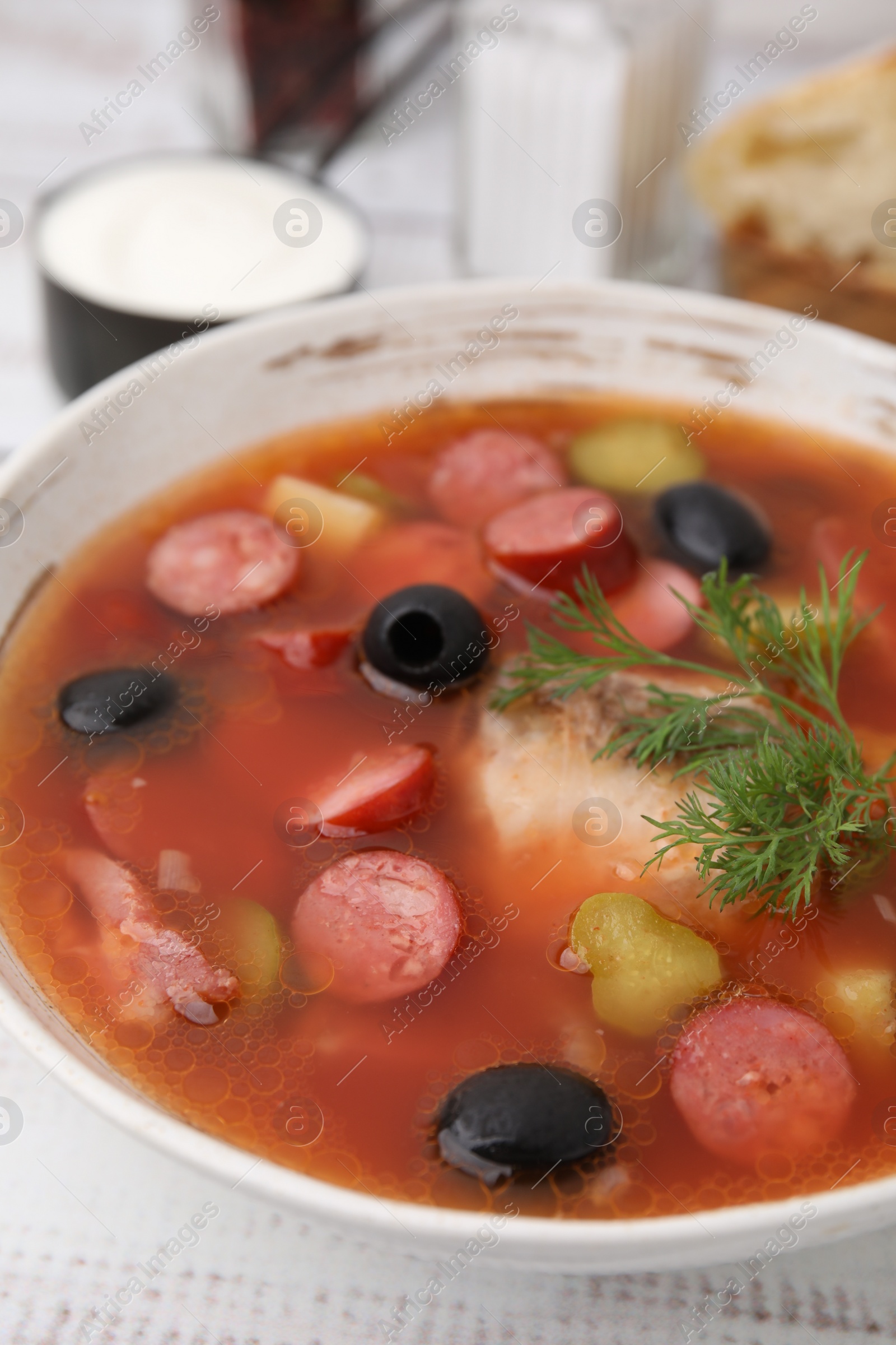 Photo of Meat solyanka soup with thin dry smoked sausages in bowl on white table, closeup